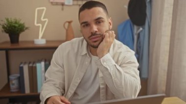 Evde dizüstü bilgisayar kullanan gülümseyen genç İspanyol adam, gündelik profesyonellik ve ulaşılabilirlik yayıyor..