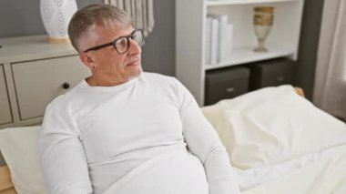 Kendine güveni tam, beyaz saçlı orta yaşlı bir adam, doğal, neşeli bir ifadeyle gülüyor, diğer tarafa bakıyor, rahat yatak odasında pijama giyiyor..