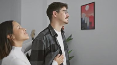 Gülümseyen bir kadın bir erkeğin koluna dokunur, modern bir oturma odasında güncel sanatla gelişigüzel giyinir..