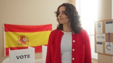 Kapalı bir üniversite odasında İspanyol bayrağı taşıyan, kırmızı hırka giyen ve oy kabininin yanında neşeli bir şekilde gülümseyen genç bir İspanyol kadın..