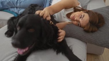 Genç bir kadın rahat bir oturma odasında dinleniyor, kanepede uzanıyor ve siyah labrador köpeğini sevgiyle okşuyor, sıcak ve samimi bir an yaşıyor..