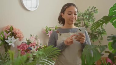 Gülümseyen genç İspanyol çiçekçi, neşe dolu çiçekçisine akıllı telefon tutarken parmaklarıyla kenarı işaret ediyor.