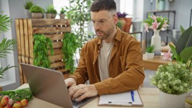 Yakışıklı İspanyol adam, yeşil ve çiçeklerle çevrili canlı bir çiçekçide dizüstü bilgisayarla çalışıyor..