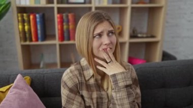 Endişeli genç sarışın kadın gergin bir şekilde evde oturuyor, sorguluyor ve endişe içinde kanepede oturuyor, elleri çenesinde.