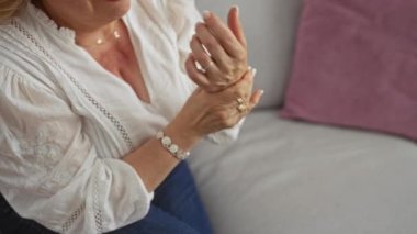 Sarı saçlı olgun bir kadın, rahat bir oturma odasında oturuyor, beyaz bir bluz ve mücevher giyerken bileğini tutuyor..