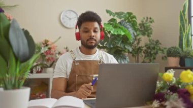 Kulaklıklı Afrikalı Amerikalı bir adam kapalı alandaki bitkilerle çevrili bir video görüşmesine katılıyor..