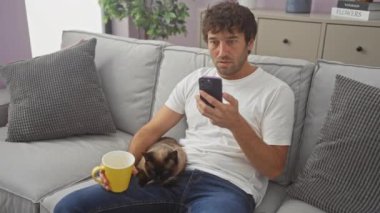 Beyaz tişörtlü bir adam evdeki kanepede oturuyor, yanında bir siyam kedisi olan sarı bir kupa ve akıllı telefon tutuyor..