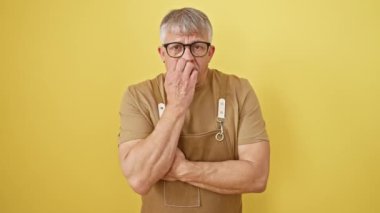 Gri saçlı, orta yaşlı, stres ve endişeyle tırnak yiyen, izole edilmiş sarı arka planda duran, önlük giyen, kendine güveni olmayan.