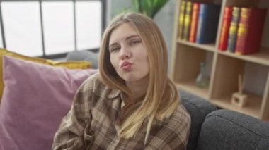 Kanepede oturan güzel sarışın kadın kameraya bir öpücük yolluyor. Seksi ve eğlenceli bir aşk ifadesi. Kapalı alan portresi..