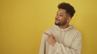 Neşeli Afro-Amerikalı adam kapüşonlu giyen, doğal, mutlu bir gülümseme gösteren. Elle yukarı, parmak yana, ifade dolu çarpıcı, izole edilmiş sarı arkaplan.