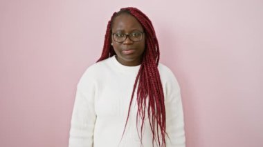 Pembe arka planda kırmızı örgülü ve gözlüklü düşünceli bir Afrikalı kadın.