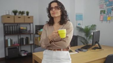 Esmer saçlı ve gözlüklü genç bir İspanyol kadın modern ofis mobilyaları ve organize raflarla bir ofiste kahve molası veriyor..
