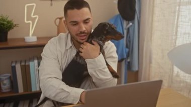 Genç bir İspanyol, rahat bir oturma odasında dizüstü bilgisayarı üzerinde çalışırken tekel köpeğine sarılıyor..