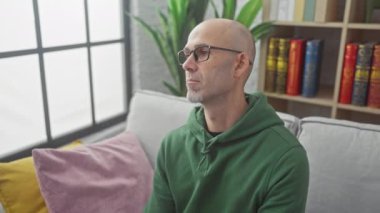 Kanepesi ve renkli yastıkları olan, gözlüklü, dalgın, kel bir adam oturma odasında dinleniyor..