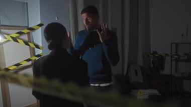 Stresli bir adam, kapalı bir suç mahallinde belli bir uyarı bandıyla bir kadın polisle konuşurken el kol hareketi yapıyor..