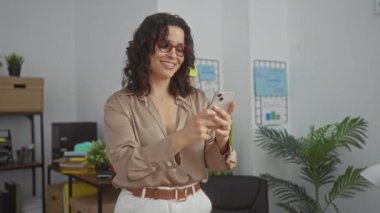 Genç, çekici esmer bir kadın bir ofiste duruyor, elinde akıllı bir telefonla gülümsüyor..