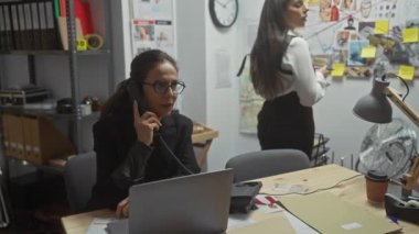 İki kadın müfettiş dağınık bir ofiste çalışıyor, biri telefonda, diğeri de kanıtları inceliyor..