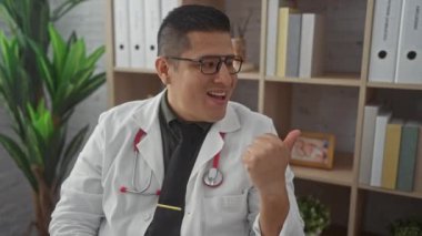 İspanyol asıllı genç adam, doktor üniforması içinde, mutlu bir şekilde yan tarafı işaret ediyor, klinikte ağzı açık gülümsüyor.