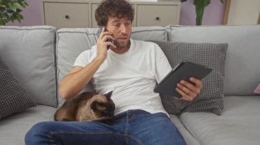 Genç bir adam evinde, tabletinde ve telefonunda, yanında rahat bir kanepede siyam kedisi olan birden fazla iş yapıyor..
