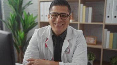 Gözlüklü, gülümseyen İspanyol erkek doktor hastane ofisinde güven içinde oturuyor, profesyonellik ve dostluk saçıyor..