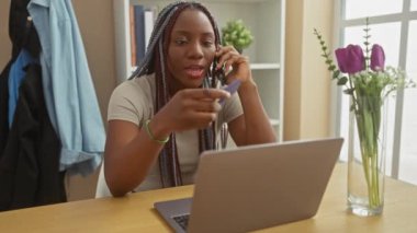 Saç örgülü Afrikalı bir kadın evde kredi kartı ve laptopuyla alışveriş yapıyor, telefonu kulağına tutuyor.