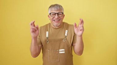 Neşeli, gri saçlı, gözlüklü ve önlüklü orta yaşlı adam umut dolu gülümsemesi ve gözleri kapalı parmaklarını çapraz yapıyor. Sarı izole edilmiş arka planda göze çarpan batıl inanç sahnesi.