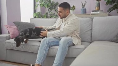 Gündelik giyinmiş genç bir İspanyol iyi döşenmiş oturma odasındaki rahat bir kanepede köpeğini sevgiyle besliyor..