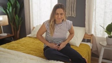 Karın ağrısı çeken genç, sarışın bir kadın evdeki rahat bir yatak odasında oturuyor..