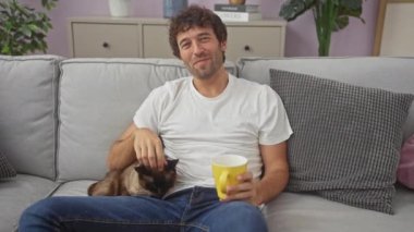 Evde bir kanepede oturan, bir kediyi okşayan ve elinde sarı bir kupa tutan rahat bir adam..
