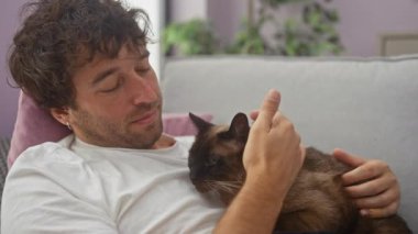 Oturma odasında genç bir İspanyol adam siyam kedisiyle sevgi dolu bir şekilde sarılıp ev içinde samimi bir bağ ve rahatlama sahnesi yaratıyor..