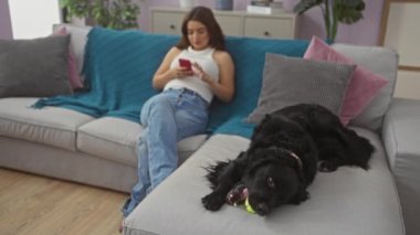 Genç bir İspanyol kadın modern bir oturma odasındaki bej bir kanepede dinleniyor. Siyah labrador köpeği yakınlarda oynarken telefonuyla mesajlaşıyor..