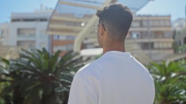 Beyaz gömlek giyen, gözlüklü genç, İspanyol bir adam arka planda palmiye ağaçları ve binaları olan bir şehir parkında dikiliyor..