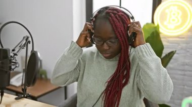 Bir müzik stüdyosunda saç örgülü podcast 'i olan, gözlüklü ve arka planda bitcoin logosu olan yeşil kazaklı Afrikalı Amerikalı bir kadın.