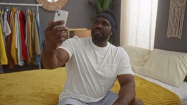 Genç Afro-Amerikalı bir adam rahat bir yatak odasında bir yatakta oturuyor, akıllı telefonuyla özçekim yapıyor, tertipli kıyafetlerin arka planına karşı..