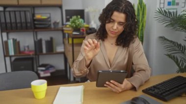 Genç esmer bir kadın ofiste çalışıyor, tablet kullanırken bir kağıda notlar yazıyor, arka planda sarı bir fincanla modern bir iş yerinde ve organize edilmiş raflar var..