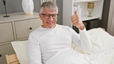 Orta yaşlı, neşeli gri saçlı bir adam pijama giyiyor, yatak odasında mutlu bir şekilde baş parmak kaldırıyor, başarıyı onaylıyor, gerçek bir kazanan gibi görünüyor.!
