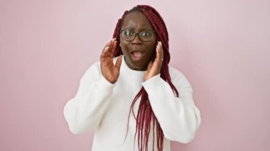 Süslü ve şaşkın bir Afrikalı kadın pembe bir arka plana karşı duruyor, heyecan ya da şok belirtileri gösteriyor, gözlük takıyor ve beyaz bir kazak giyiyor..