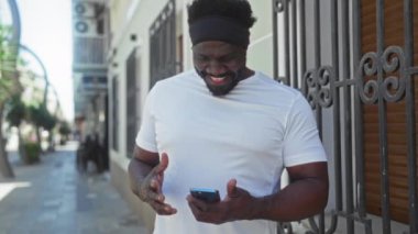 Beyaz gömlekli yakışıklı Afro-Amerikalı adam güneşli bir şehir sokağında telefonuna bakarken gülümsüyor ve sevinçle tepki veriyor..