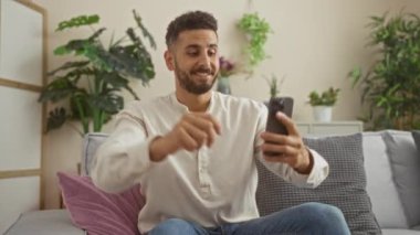 Genç İspanyol bir adam, oturma odasındaki kanepede içi bitki ve rahat yastıklarla otururken kollarını kaldırarak kutlama yapıyor..