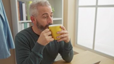 Ev ortamında sakallı, orta yaşlı bir adam sarı bardaktan kahvenin tadını çıkarıyor..