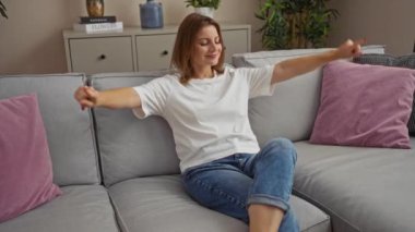 Genç, çekici bir kadın oturma odasındaki rahat bir kanepede kollarını açmış rahat bir ev ortamında rahatlık ve rahatlık hissi veriyor..