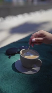 Bir kadın turkuaz kumaşla kahve karıştırıyor güneş gözlükleriyle, rahatlatıcı bir açık hava anı yakalıyor..