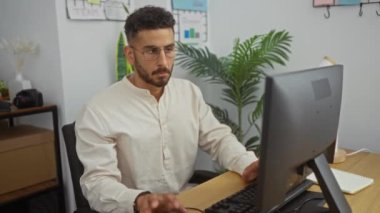 Ofiste çalışan, gözlüklü, odaklanmış ve kendini işine adamış, bilgisayar ekranı ve arka planda bitkileri olan yakışıklı İspanyol bir adam..