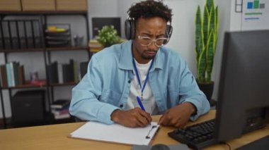 Ofiste gözlüklü, kulaklık takan, bilgisayarla uğraşırken kalemle notlar yazan Afrikalı genç bir Amerikalı..