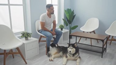 Hispanik bir adam veteriner kliniğinde bir kurt köpeğiyle düşünceli bir şekilde oturuyor.