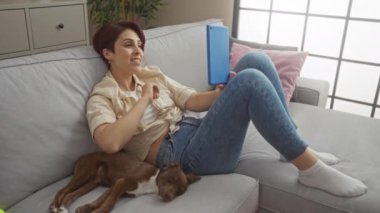 Köpeğiyle oturma odasında dinlenen genç bir kadın, modern bir dairede gri bir kanepede uzanırken mavi bir tablet kullanarak bir video konferansa katılıyor..