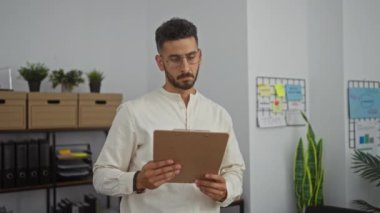 Ofiste elinde pano olan İspanyol bir adam. Etrafı bitkilerle, ofis malzemeleriyle ve duvarlarda organizasyonel çizelgelerle çevrili..