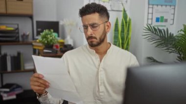 Ofisteki genç İspanyol adam, bitki ve raflarla çevrili bir belgeyi içeride okumak için gözlüklerini çıkarıyor..