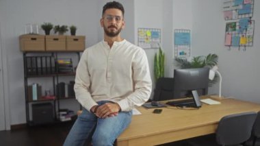 Gündelik beyaz gömlekli çekici bir genç adam modern bir ofiste masanın üzerinde oturuyor. Kollarını kavuşturup kameraya gülümsüyor..