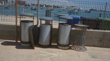 Güneşli bir günde arka planda deniz ve teknelerin olduğu Porto Sezaryo, İtalya 'daki bir rıhtımın yakınındaki paslı metal çöp kutuları..